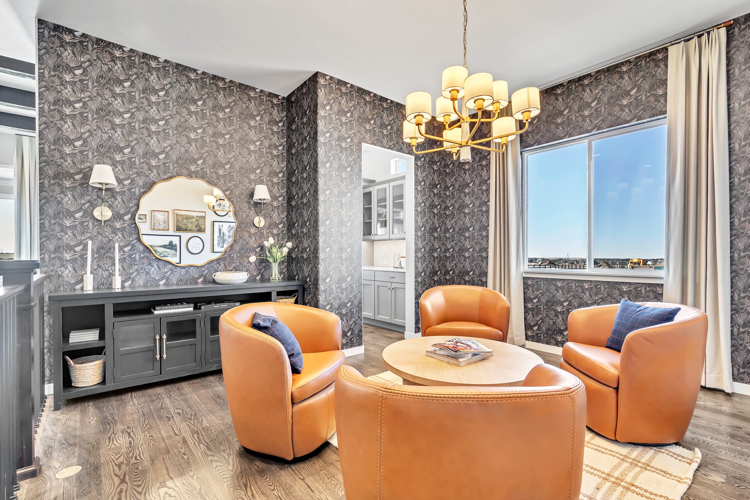 Modern sitting area featuring four caramel-colored leather swivel chairs arranged around a light wood round table, set on a patterned rug. A large window with cream curtains lets in natural light, highlighting the dark floral wallpaper. A black console table against the wall is decorated with a scalloped-edge mirror, framed art, white sconces, candles, and a vase of flowers. A gold chandelier with white shades hangs from the ceiling, adding a touch of elegance