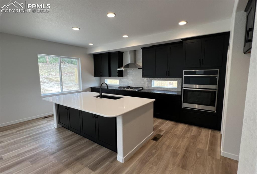 Kitchen featuring dark cabinetry, recessed lighting, wall chimney exhaust hood, a center island with sink, and light wood-style flooring